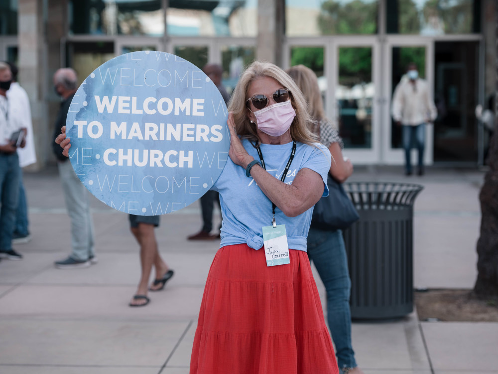 Person with a mask holding a circular sign that states 'Welcome to Mariners Church'